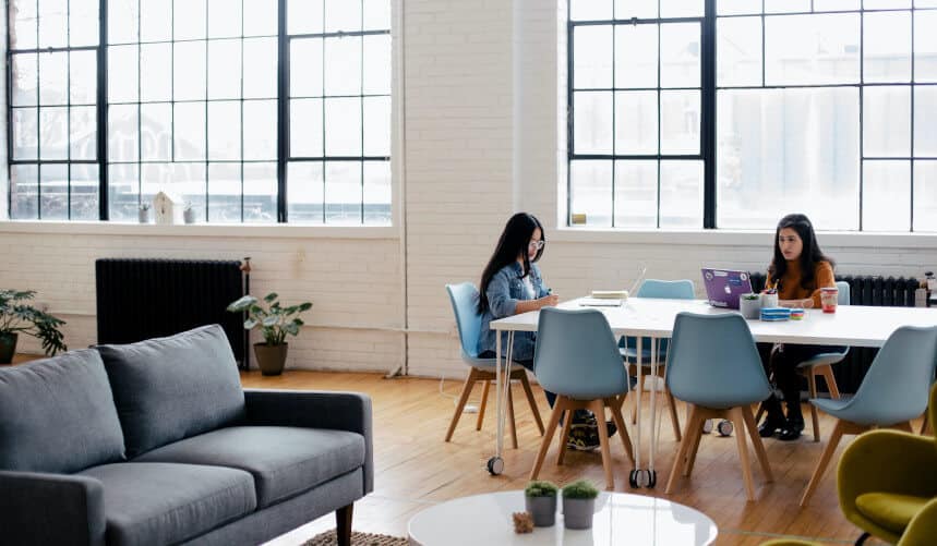Two people work at a table in a bright room with large windows, modern furniture, and plants.