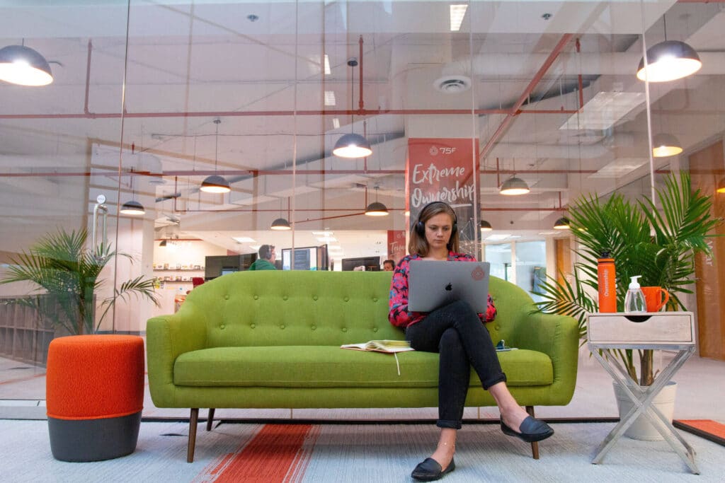 Person sitting on a green couch in a modern office with glass walls, using a laptop.