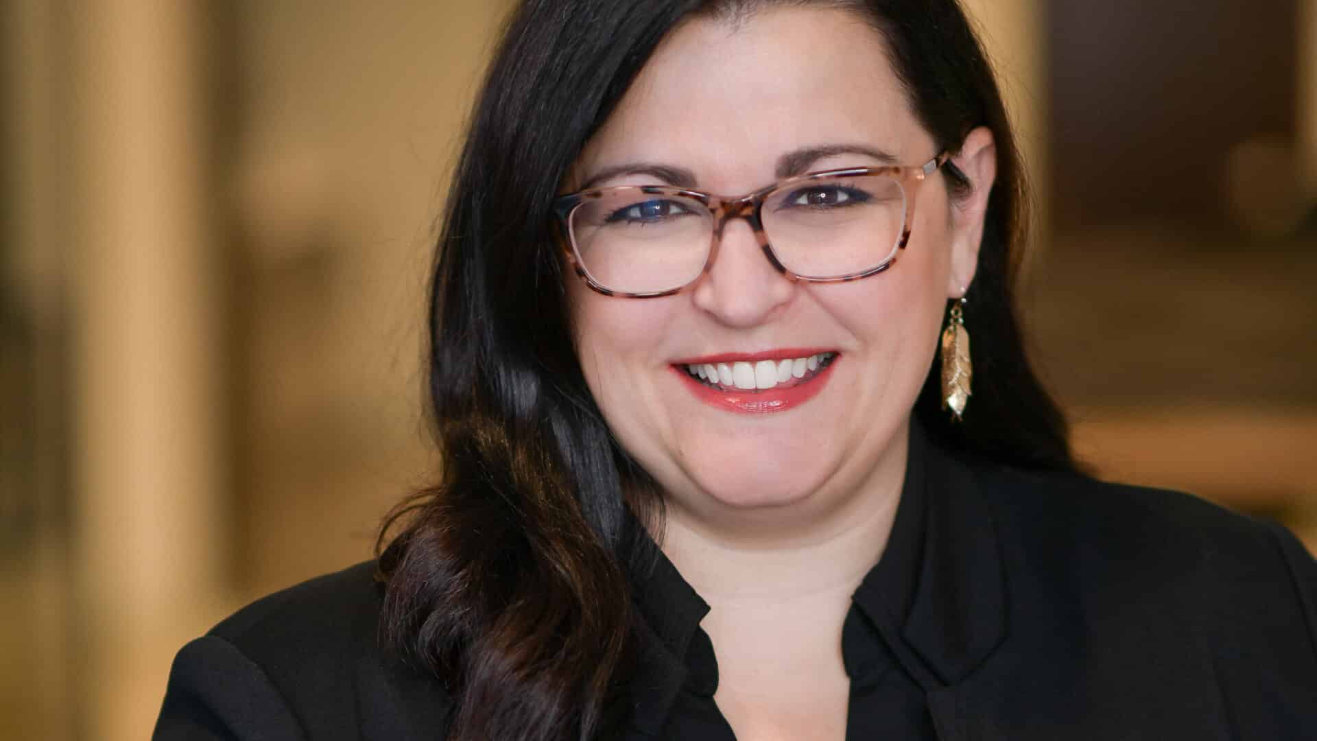 Person with long dark hair and glasses smiling, wearing a black top and earrings. Blurred neutral background.