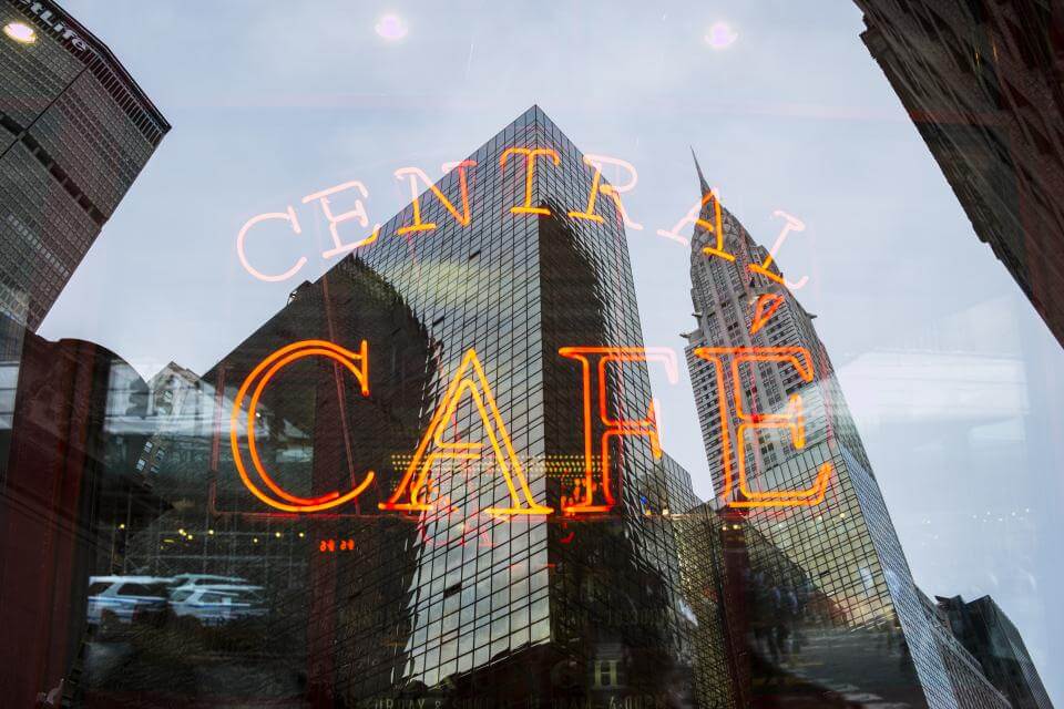 Neon "Central Café" sign reflected in glass, with skyscrapers and buildings visible in the background.