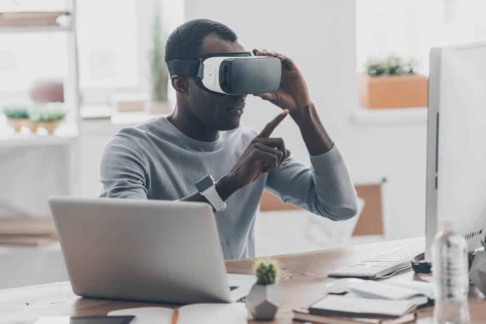 Person using a VR headset at a desk with a laptop, pointing at a computer monitor in a bright office.