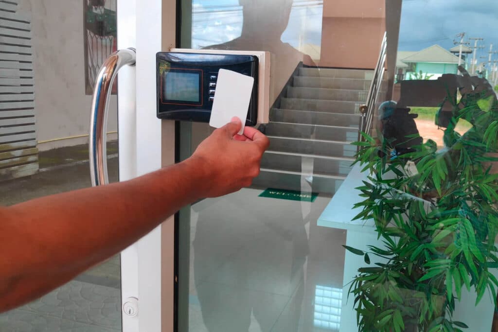 A person holding a card near a digital access reader by a glass door, with stairs visible in the background.