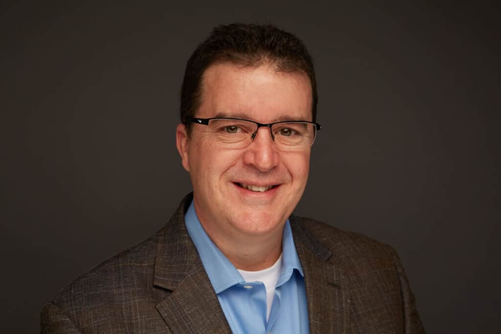 Man wearing glasses, smiling, in a blue shirt and brown blazer against a dark background.