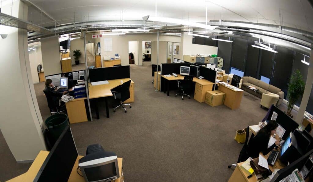 Wide-angle view of an open-plan office with desks, chairs, computers, and boxes; few people are working.