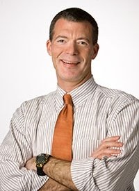 Man in a striped shirt with an orange tie, smiling, arms crossed, against a plain white background.