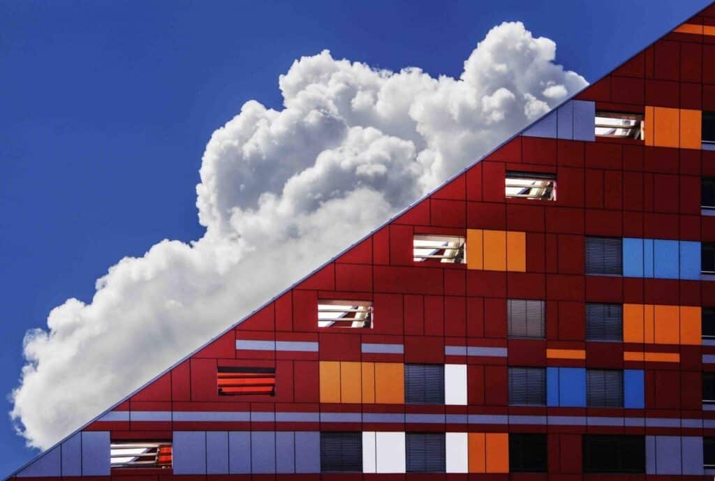 Modern red building with colorful geometric panels angled against a backdrop of a bright blue sky and fluffy clouds.