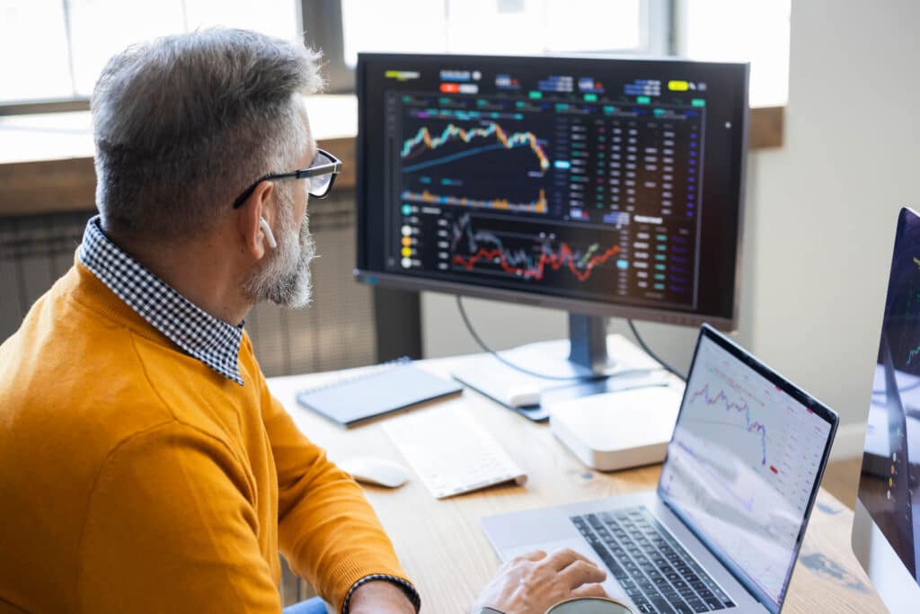 Person in orange sweater analyzing stock charts on a computer and laptop in an office setting.