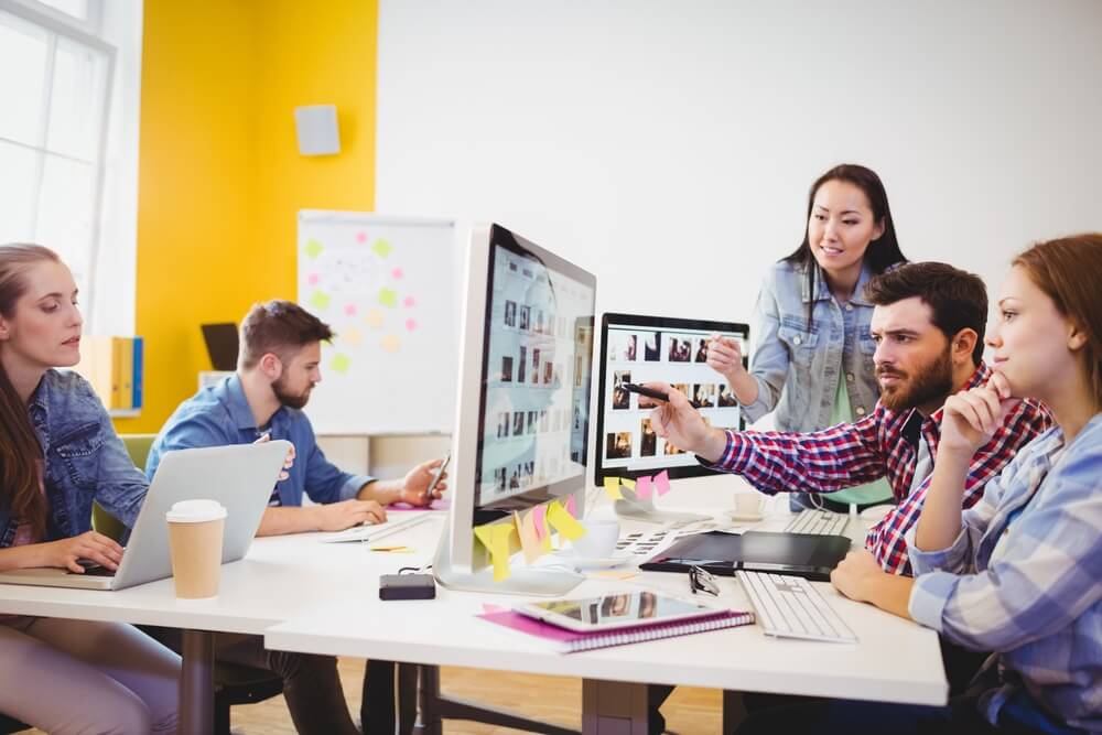 Five people in a bright office collaborate at desks with computers, discussing and working on a project.
