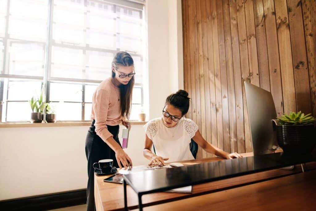 Two women working together at a desk with papers, in an office with wooden walls and potted plants.