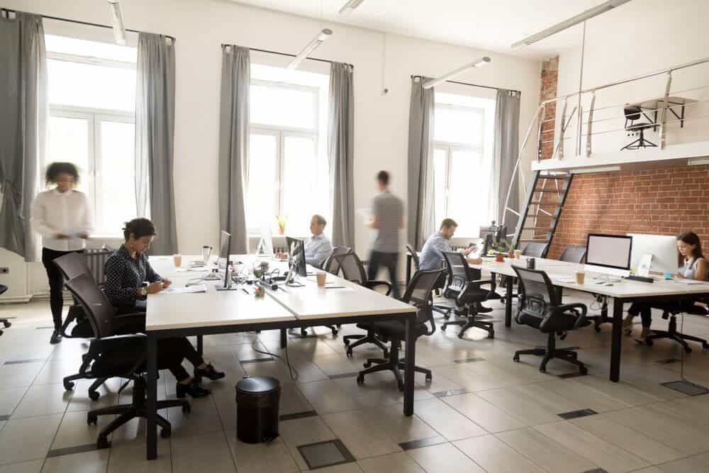 Open office with people working at desks, laptops open, large windows, curtains, and a mezzanine with stairs.