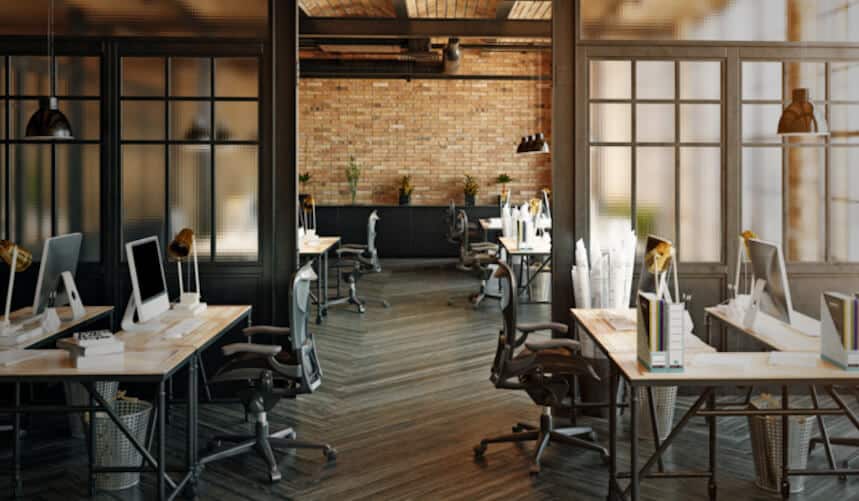 Modern open office with wooden floors, computer desks, chairs, and exposed brick walls.