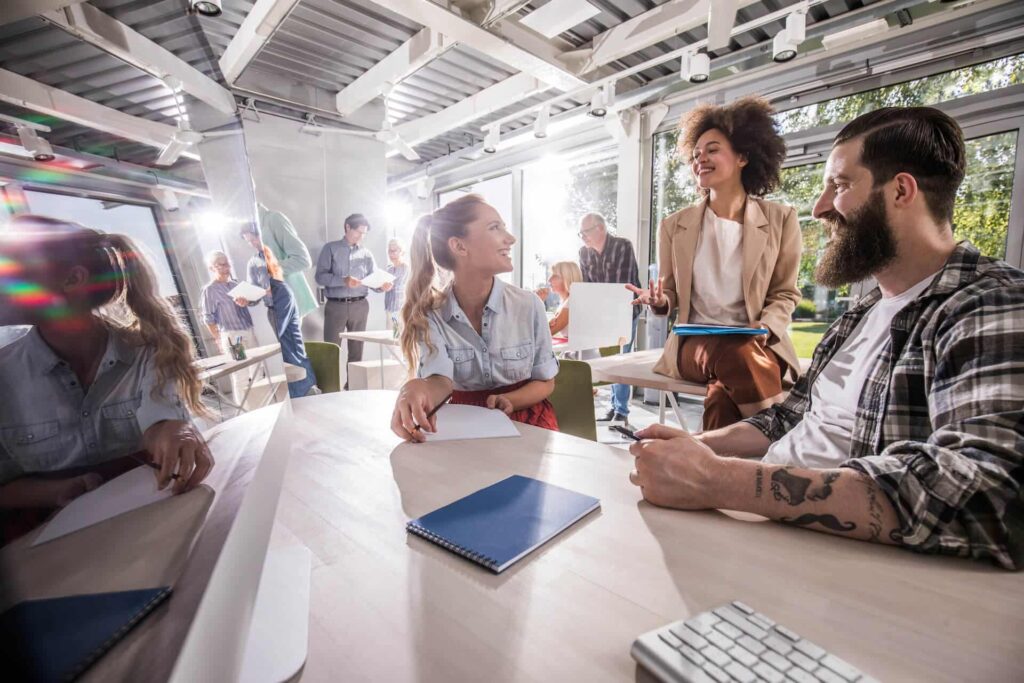 A diverse group of people engaged in a lively discussion around a table in a sunlit modern office space.