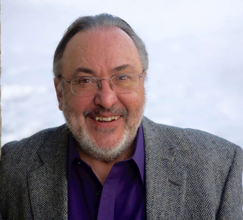 Smiling man with glasses, wearing a purple shirt and gray herringbone jacket, against a light background.