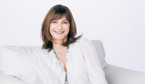 Smiling woman with brown hair in a white sweater, sitting on a white couch against a plain background.