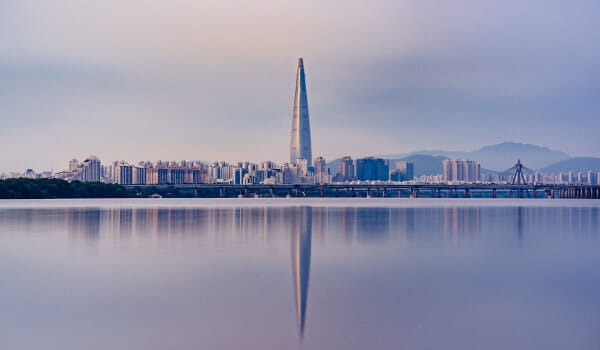 City skyline with a tall tower reflected in a calm river at dusk, mountains in the background.