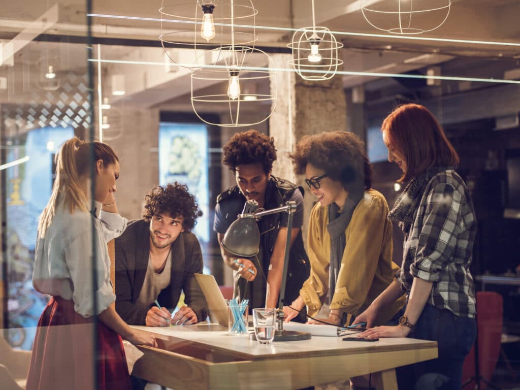 A group of people collaborating around a table in a modern office with hanging lights.