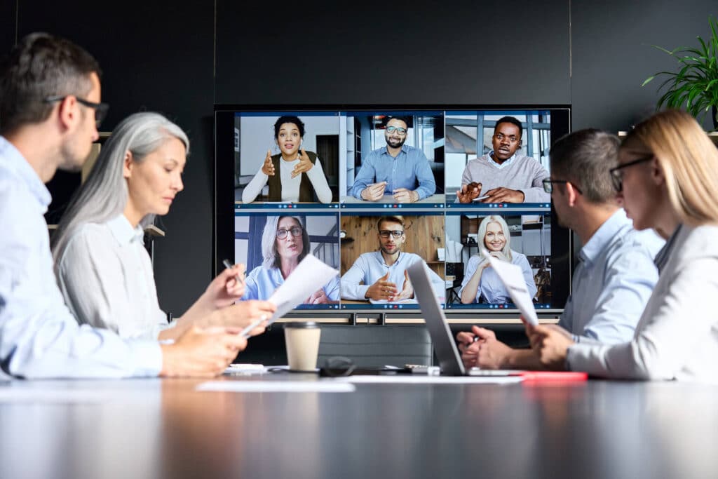 People in a conference room video-calling colleagues displayed on a large screen, engaging in a virtual meeting.