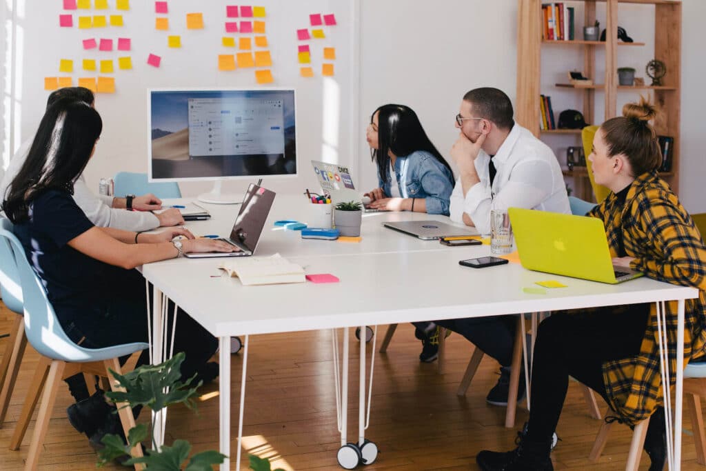 Five people in a meeting room, working on laptops, with sticky notes on the wall and a large monitor displaying content.