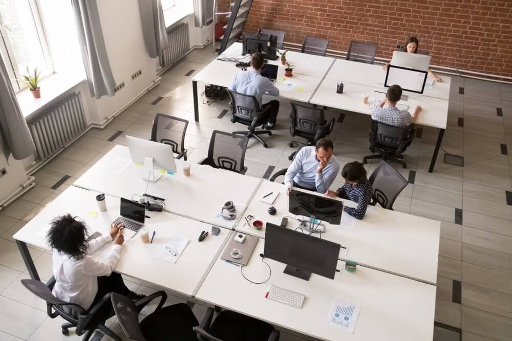 People working at desks in an open office space with computers, plants, and a brick wall.