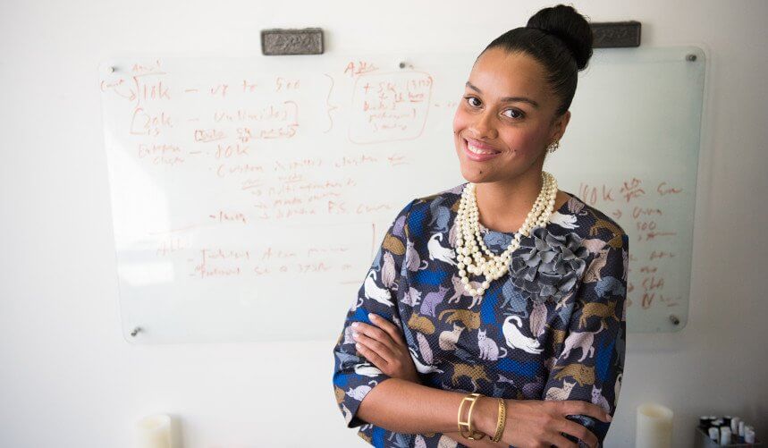 Smiling woman in patterned dress stands confidently with arms crossed in front of a whiteboard with writing.