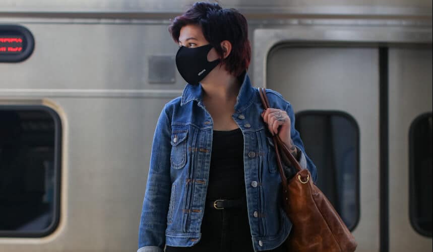 Person in denim jacket and black mask stands on train platform, holding a brown bag.