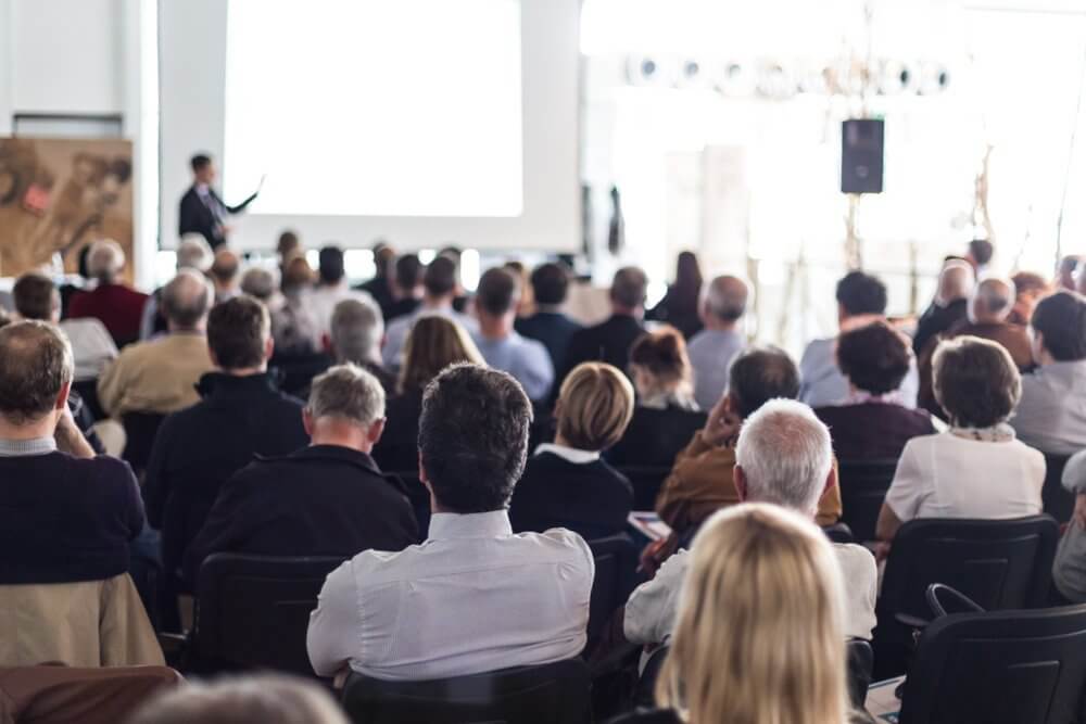 Audience attending a presentation in a conference room with a speaker and large screen.