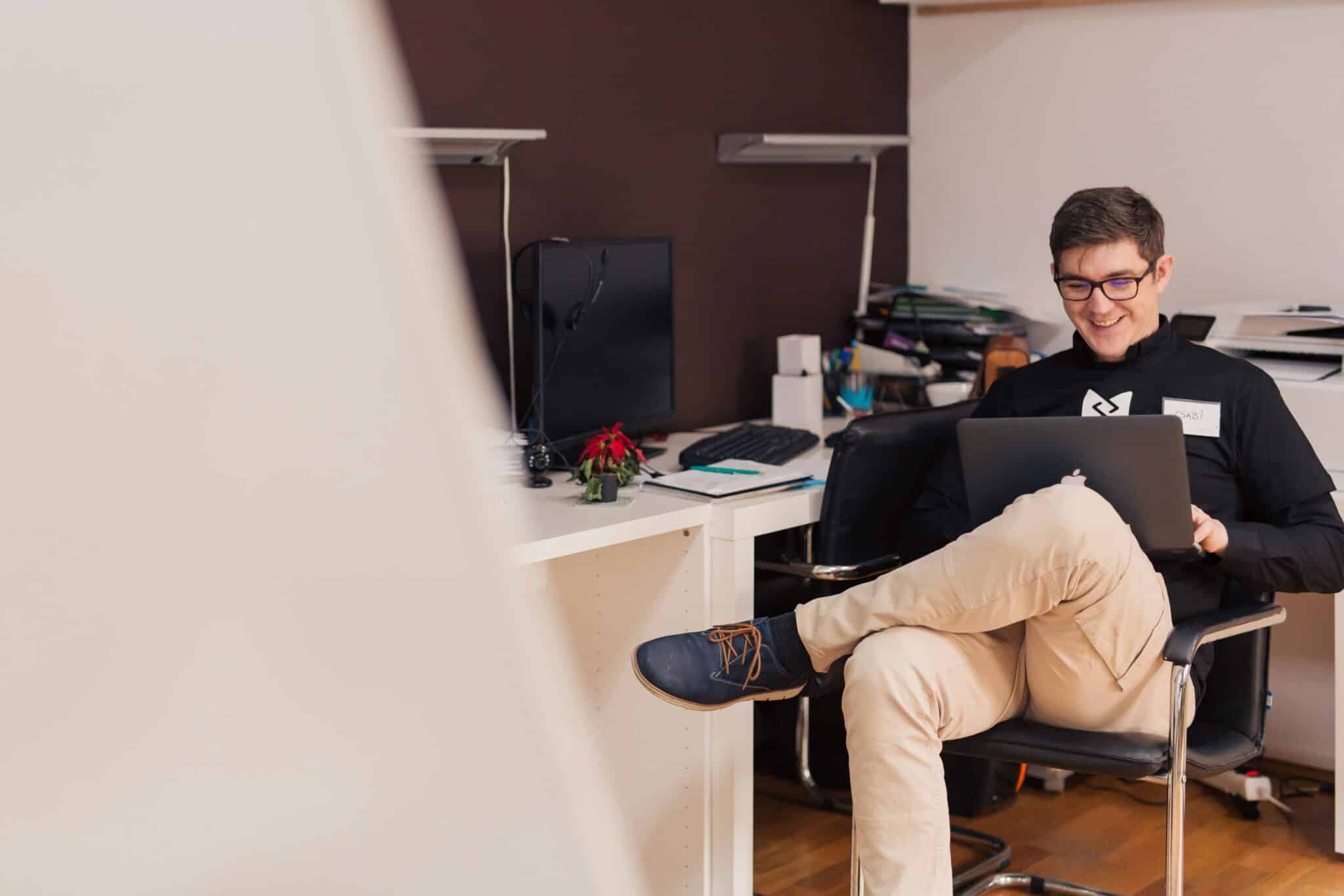Person sitting in an office chair, smiling at a laptop, with a cluttered desk in the background.