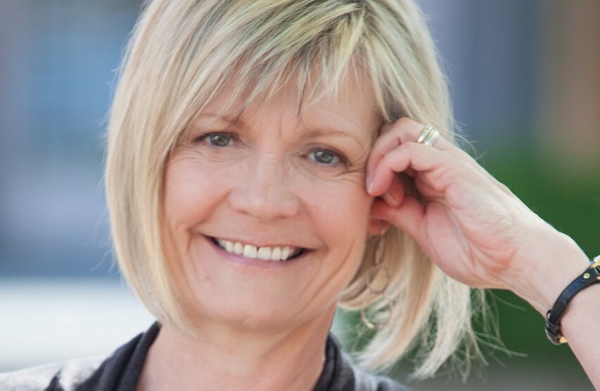 Smiling woman with short blonde hair, wearing a black top, rests her hand on her face. Blurred background.
