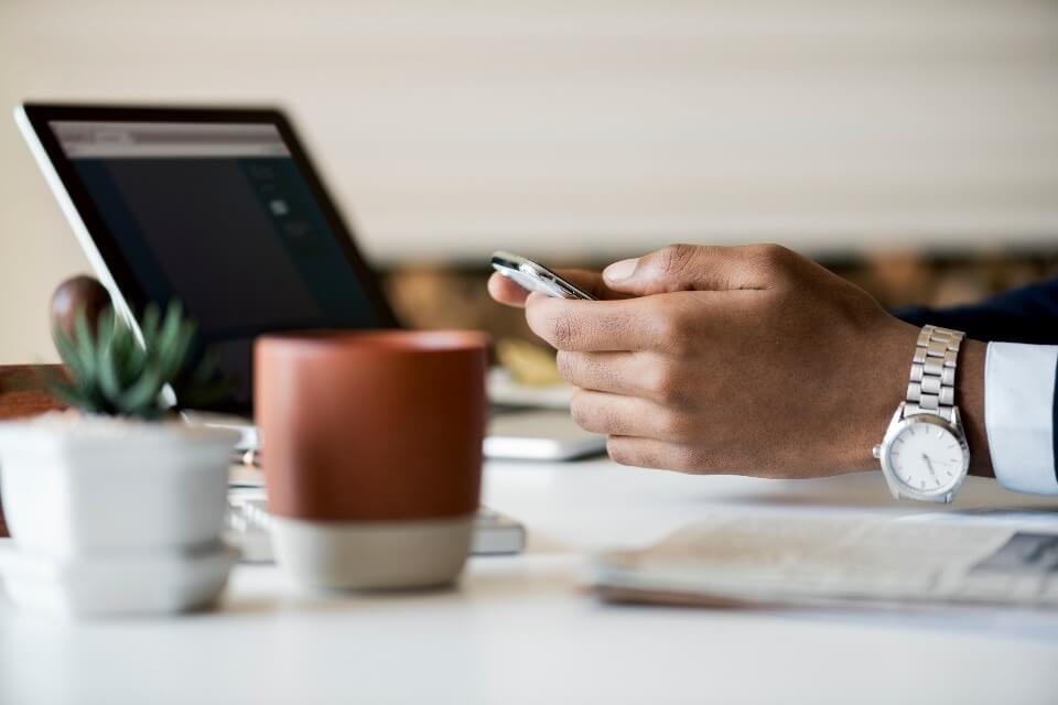 Person holding a smartphone at a desk with a laptop, plant, and coffee cup.