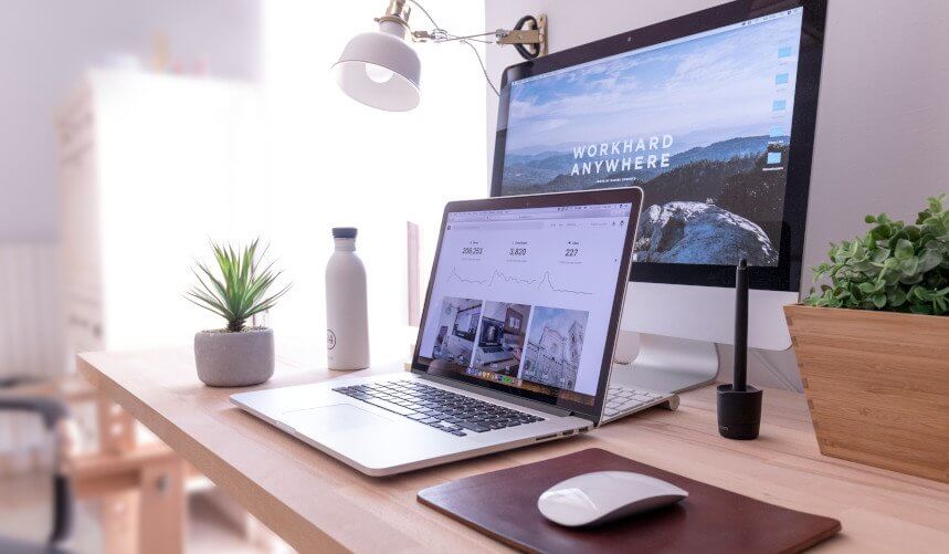 A workspace with a laptop, desktop monitor, plant, water bottle, and mouse on a wooden desk.