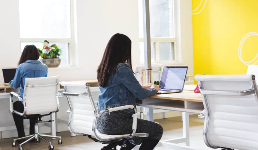 Two people working on laptops at a long desk in a bright, modern office with white chairs and a yellow accent wall.