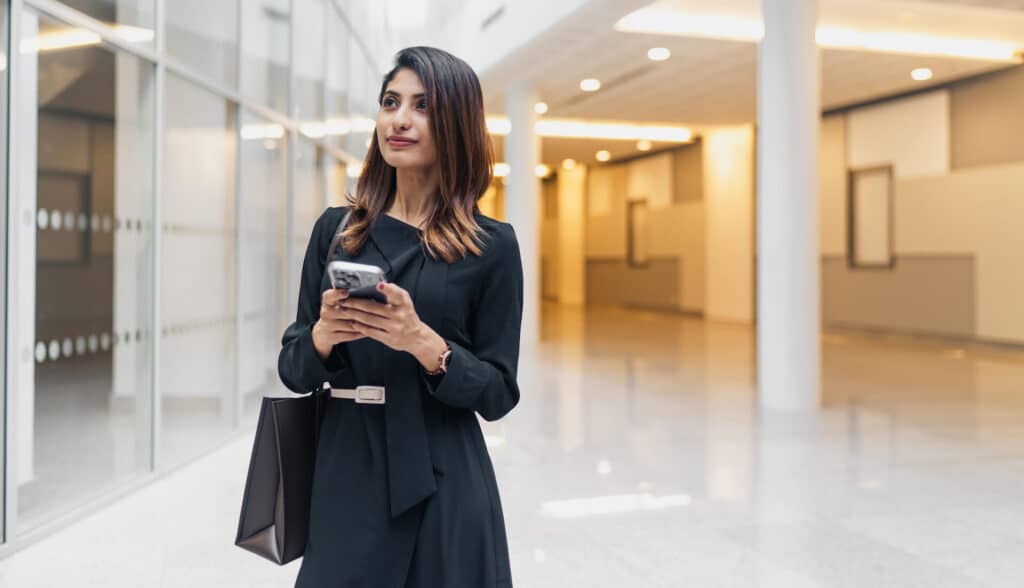 Woman in black dress holding a phone and bag, walking through a modern, brightly lit hallway.