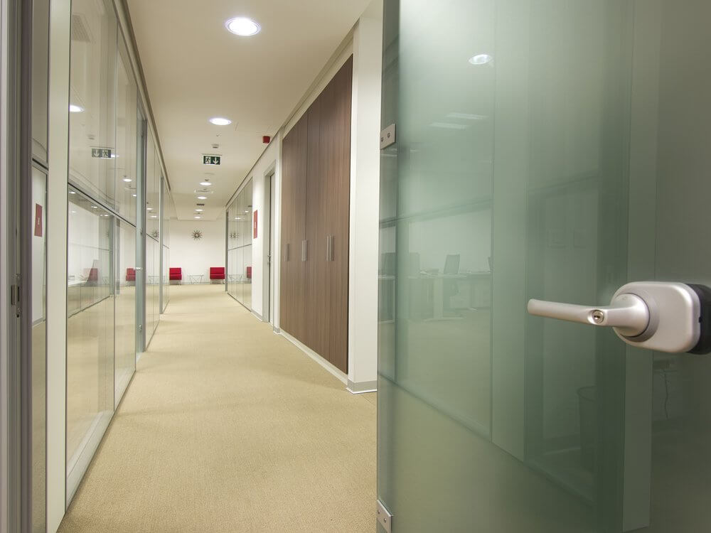Open office door leading to a hallway with glass partitions, beige carpet, and red chairs in the distance.