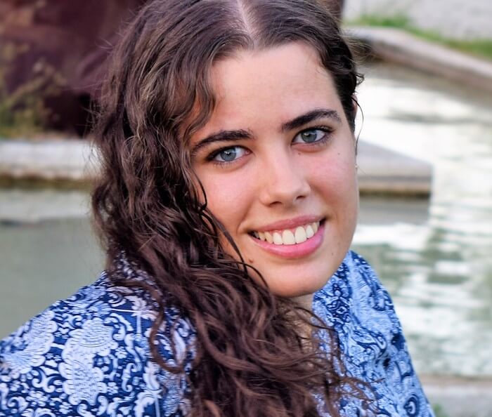 Person with long curly hair and blue patterned shirt smiling near water.