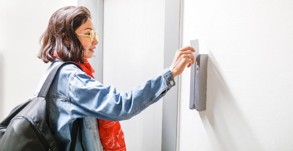 Woman with a backpack uses a key card to access a building, wearing sunglasses, a red scarf, and a denim jacket.