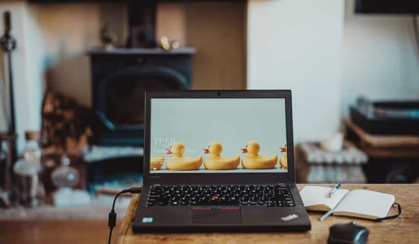 A laptop on a wooden table, displaying a wallpaper with rubber ducks. A notepad and camera are beside it.