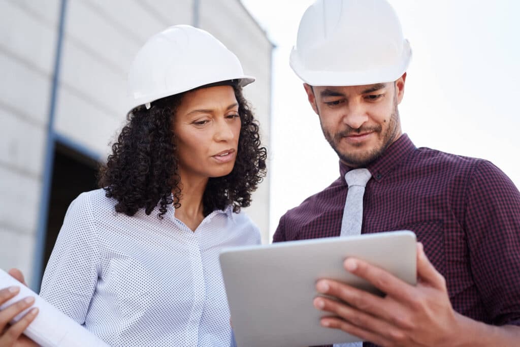 Two engineers in hard hats reviewing plans on a tablet at a construction site.