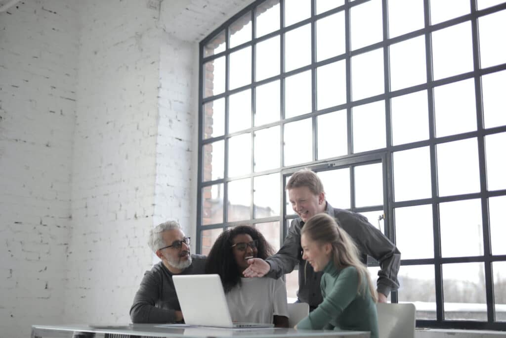 A group of four people laughing and talking around a laptop in a bright room with large windows.