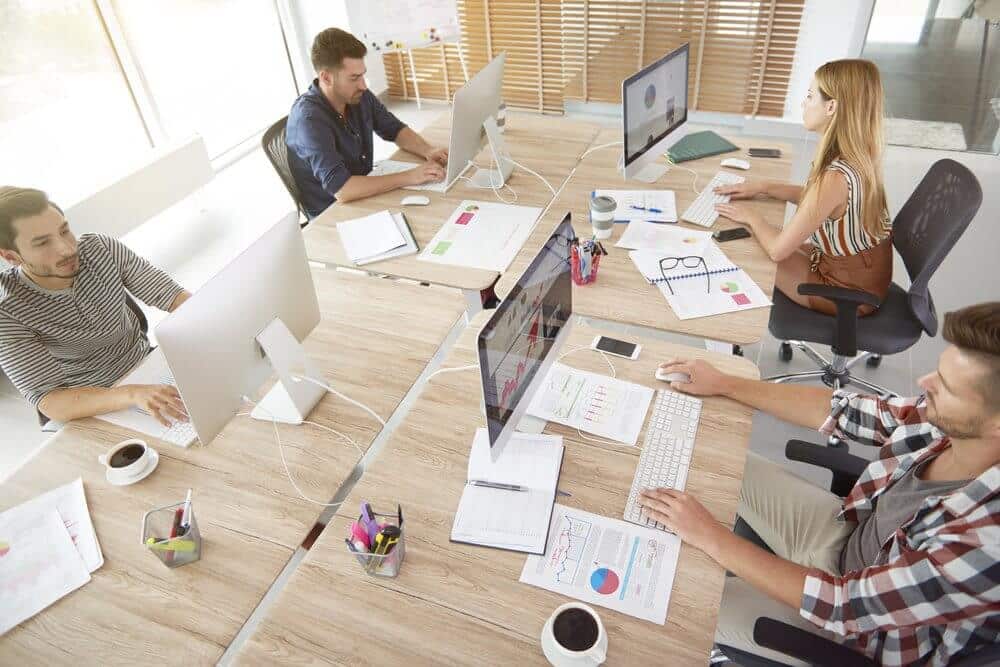 Four people working at desks with computers in a bright office, surrounded by documents and coffee cups.
