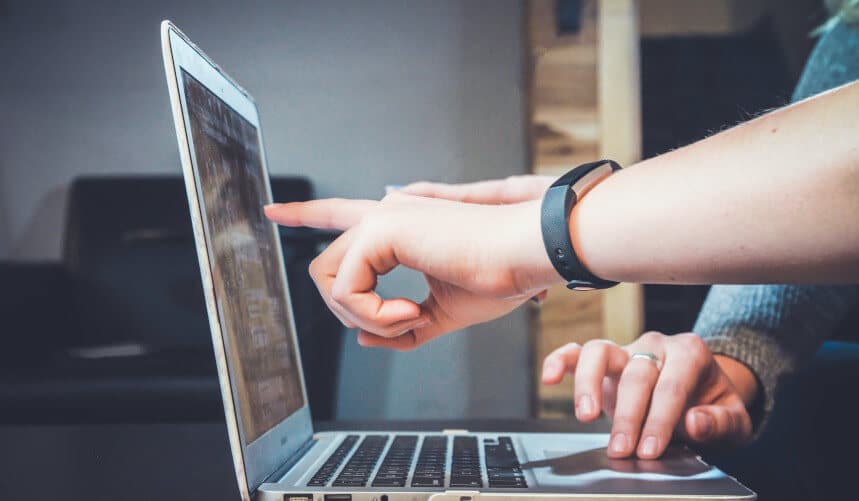 Two hands, one with a smartwatch, point and touch a laptop screen in a room with a blurred background.