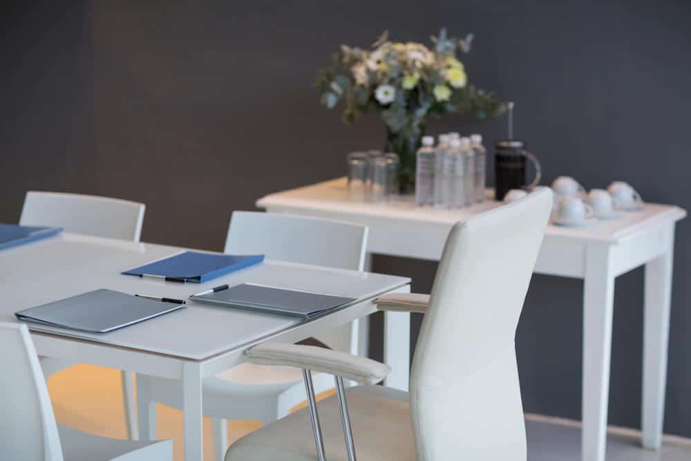 Modern conference room with a white table, notepads, and chairs. A side table holds water bottles and flowers.