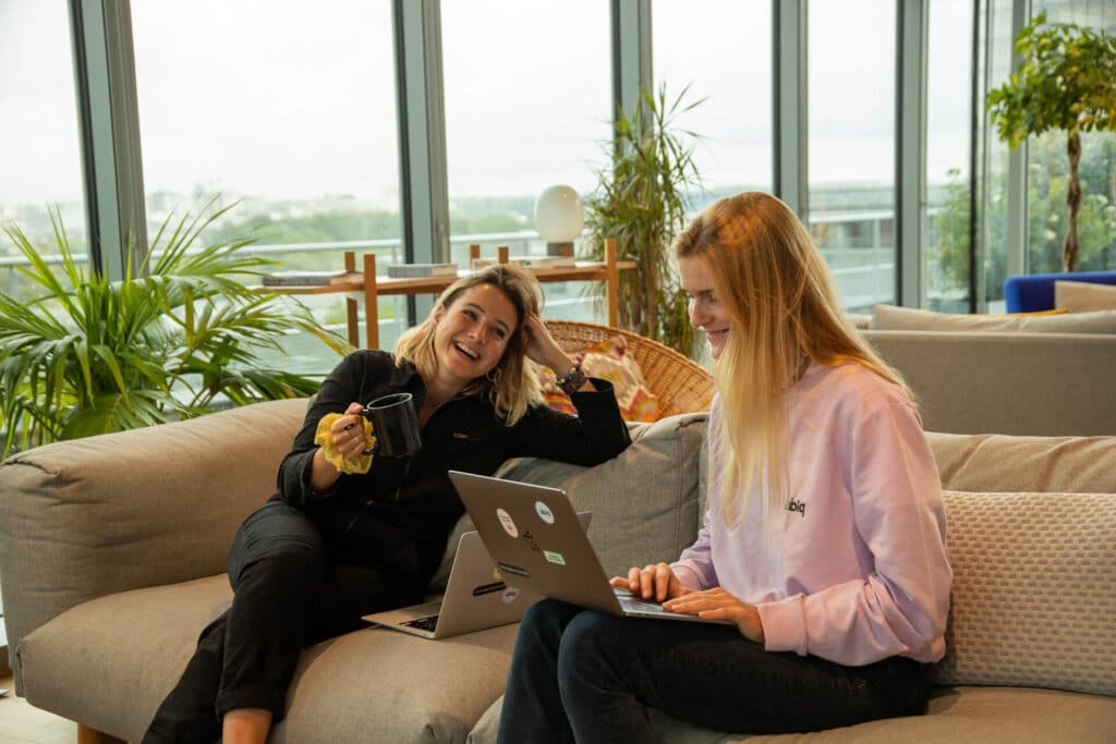 Two people sit on a couch with laptops, smiling and talking. One holds a mug. Green plants in the background.