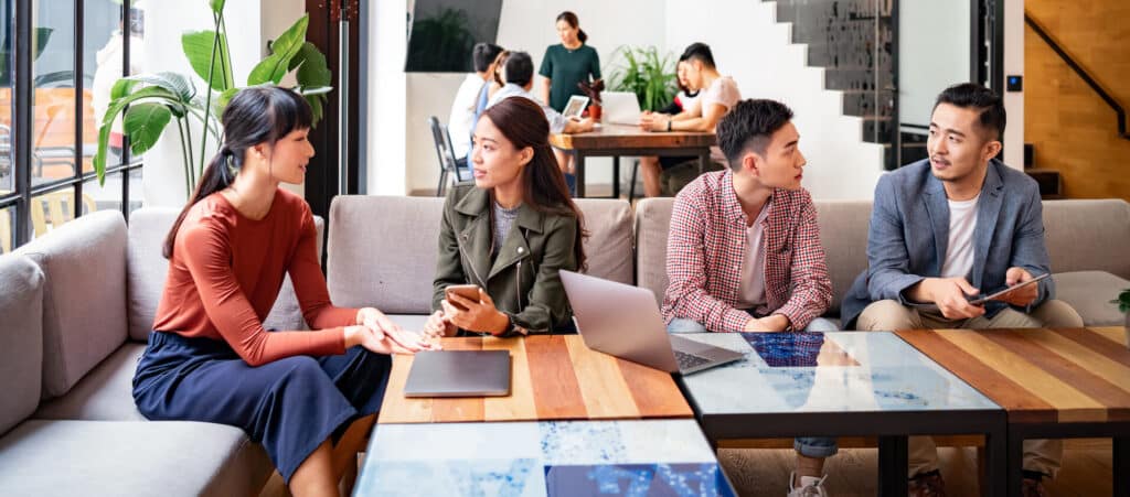 Four people discussing around a table in an office, with laptops and tablets, and others working in the background.
