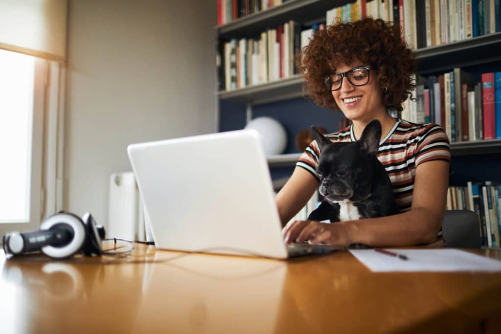 Smiling woman using a laptop with a French bulldog on her lap in a home office.