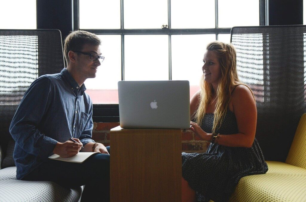 Two people seated in office, discussing work in front of a laptop, with a large window in the background.