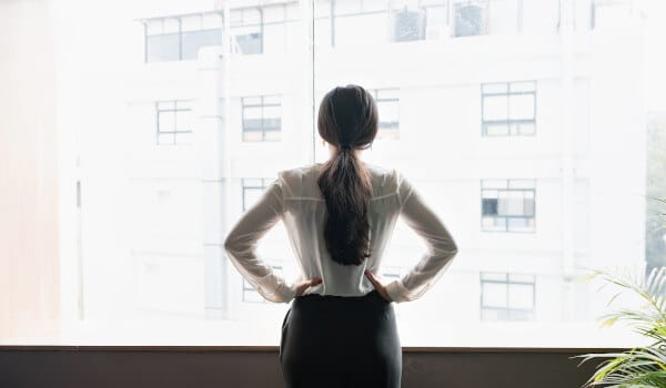 A woman with long hair stands confidently, looking out a window at a bright cityscape.