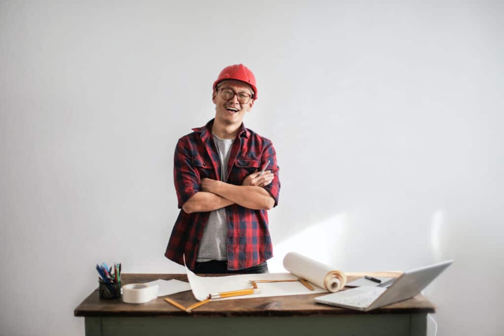 A man in a red hard hat stands smiling with arms crossed, near a desk with papers, pencils, and a laptop.