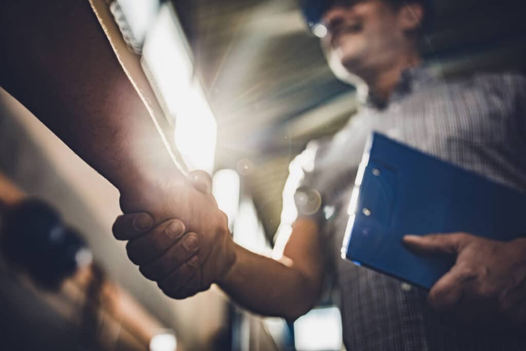 Two people shaking hands in a sunlit room, one holding a clipboard, both wearing casual shirts.