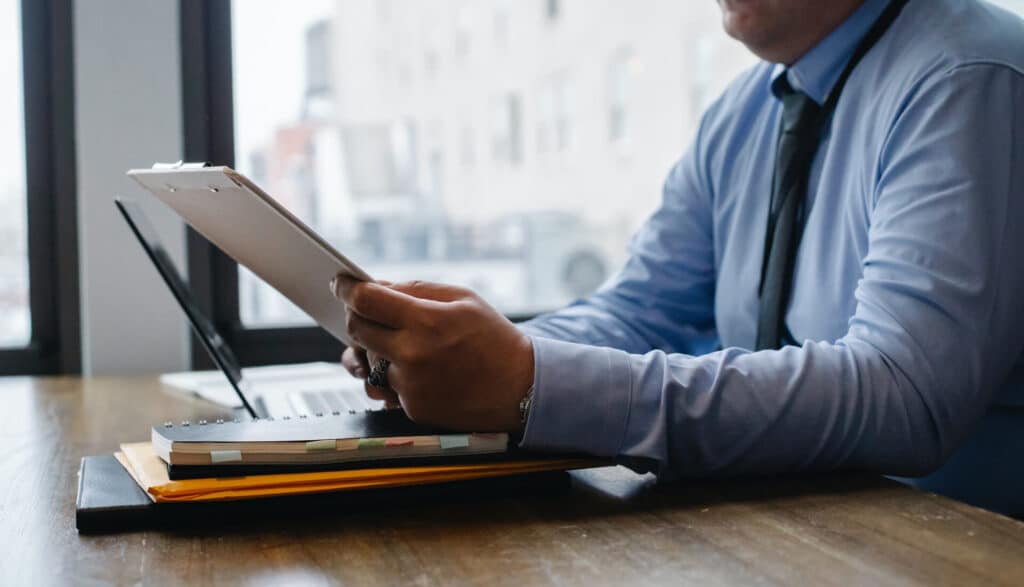 Person in a blue shirt reviews documents on a clipboard at a desk with a laptop and folders.