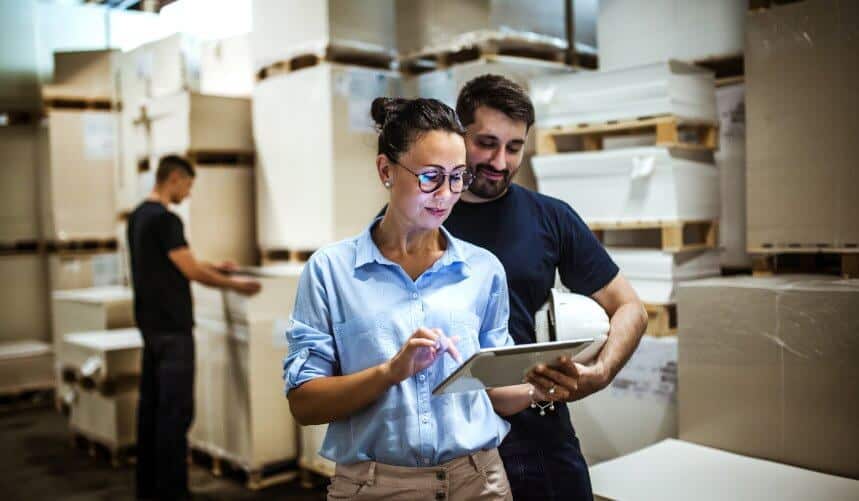 Two people in a warehouse setting looking at a tablet, surrounded by stacked pallets and boxes.
